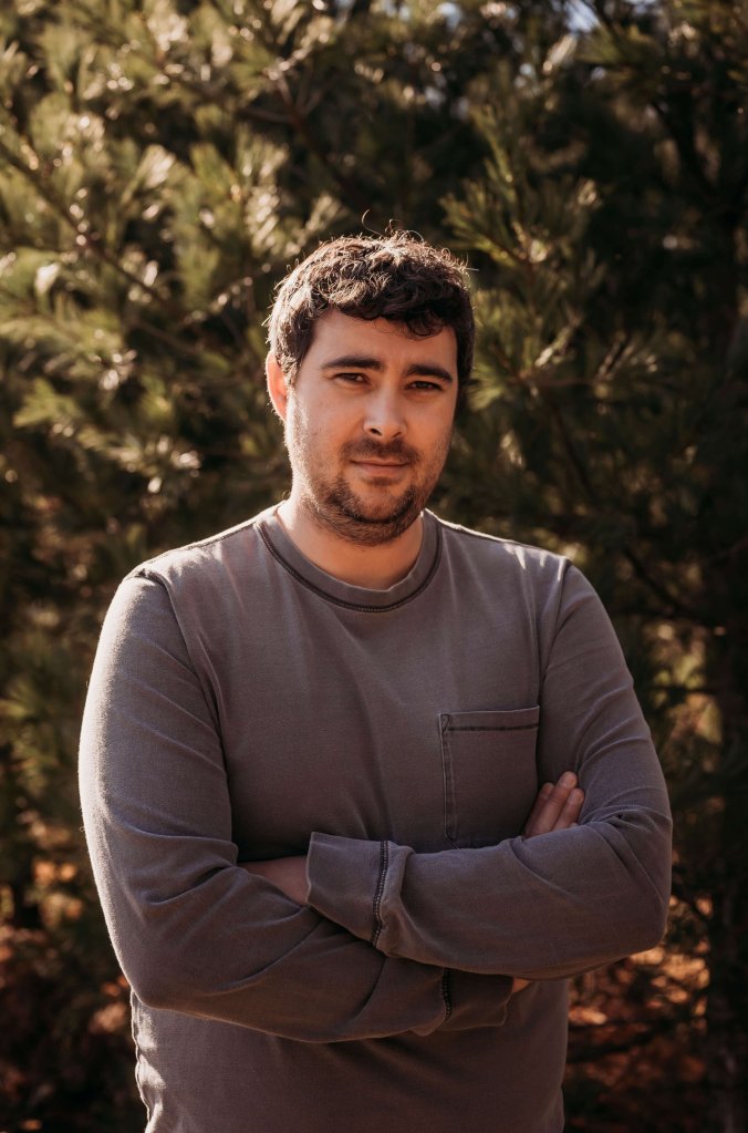 Morgan Talty: man with mustache and goatee, slightly smiling and wearing a gray long-sleeve shirt, stands with his arms crossed,  against a background of trees. 